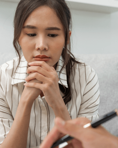 Young Asian woman leaning forward and listening to a therapist whose hand only is visible for notetaking